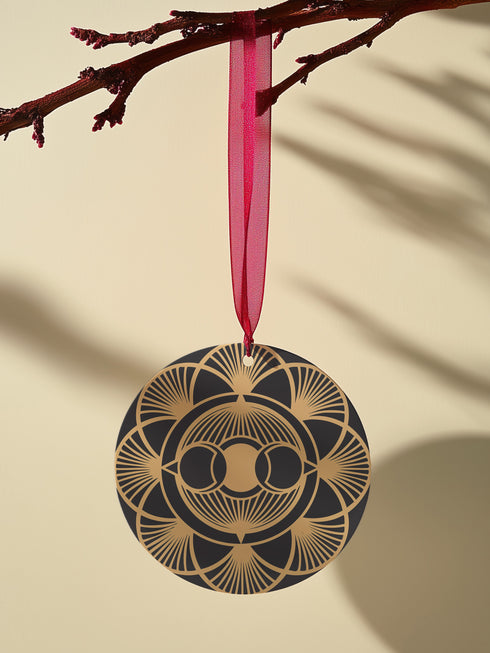 Round metal ornament featuring golden triple moon symbol surrounded by Art Deco sunburst pattern on black background. 3.5-inch diameter piece with symmetrical lunar design and crimson ribbon. Detailed celestial artwork adorns both side